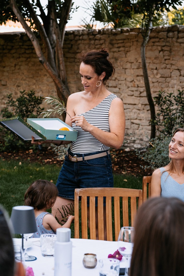 Marie-Astrid présentant une boîte à des convives attablés dans un jardin